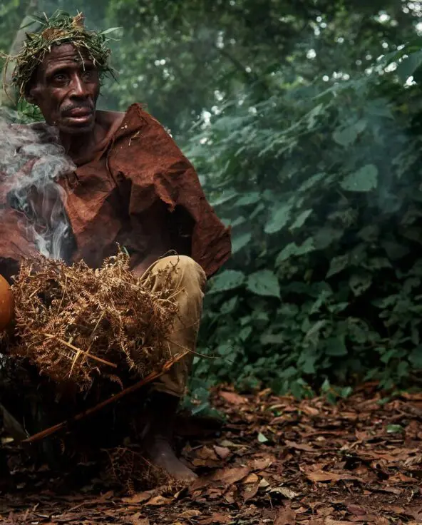 An elderly Batwa man holding a gourd and herbs, seated in a forest with smoke rising around him