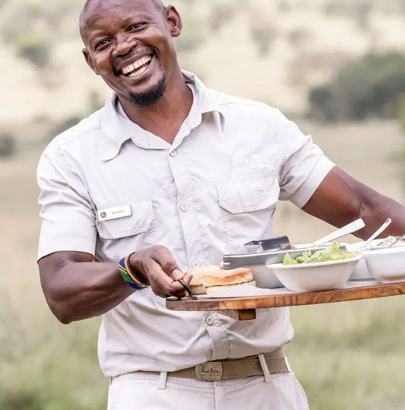 male waiter walking towards the camera smiling, serving guests lunch at olakira migration camp, tanzania, asilia africa