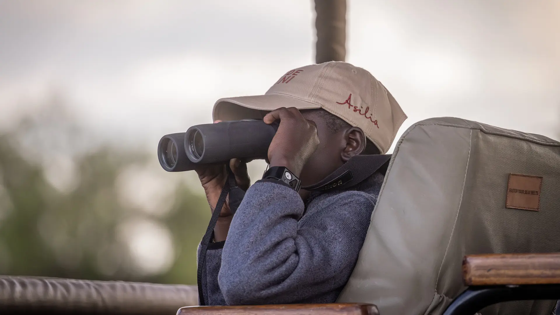 Twende Porini student holding binoculars wearing a beige Asilia cap