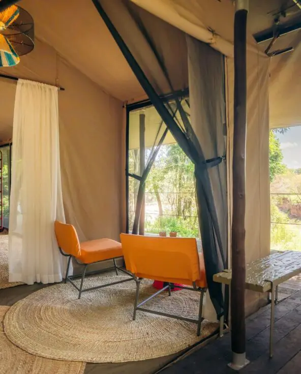 Spacious luxury safari tent at Emboo with bed, seating area, woven rugs, open canvas walls, and a private wooden deck overlooking a green riverbank landscape.