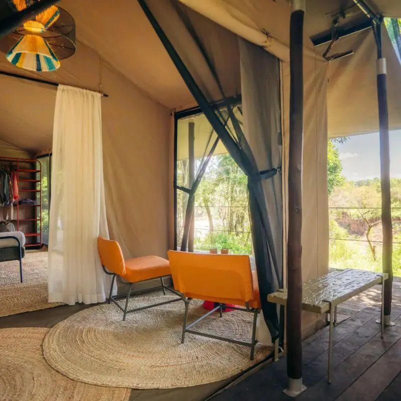 Spacious luxury safari tent at Emboo with bed, seating area, woven rugs, open canvas walls, and a private wooden deck overlooking a green riverbank landscape.