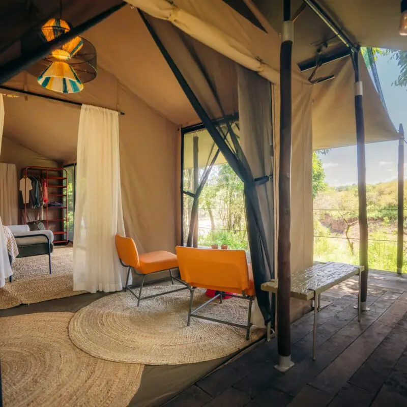 Spacious luxury safari tent at Emboo with bed, seating area, woven rugs, open canvas walls, and a private wooden deck overlooking a green riverbank landscape.