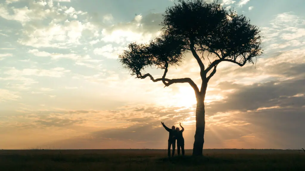 two emboo guests stand together under a tree at sunset in the maasai mara
