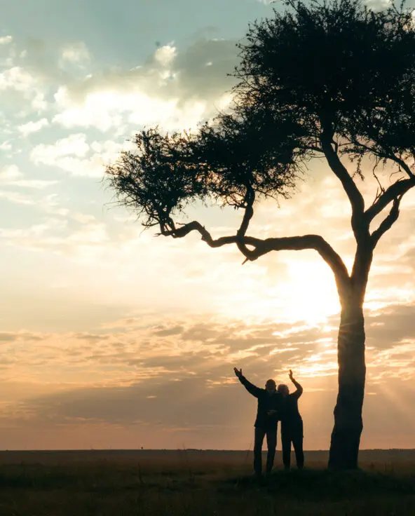 two emboo guests stand together under a tree at sunset in the maasai mara