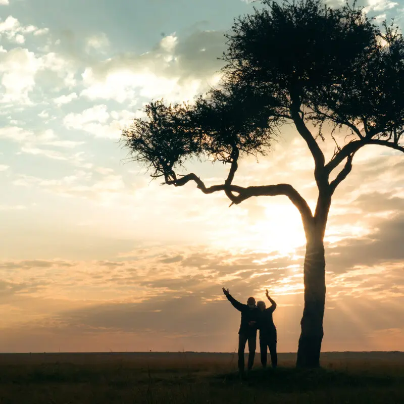 two emboo guests stand together under a tree at sunset in the maasai mara