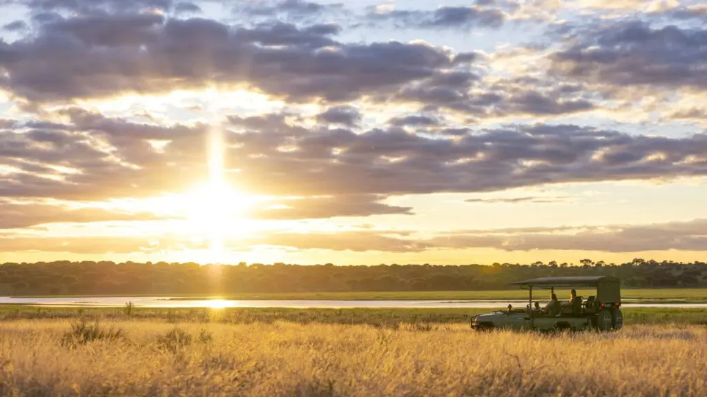 Oliver's Camp, Tarangire, an open safari vehicle carrying guests pauses in tall golden grass at sunset, overlooking a calm waterway as dramatic clouds and warm sky light