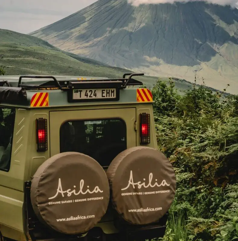 An Asilia safari vehicle parked on a green track with spare wheels visible, facing the steep volcanic slopes of Ol Doinyo Lengai rising under low clouds in the Ngorongoro Highlands.