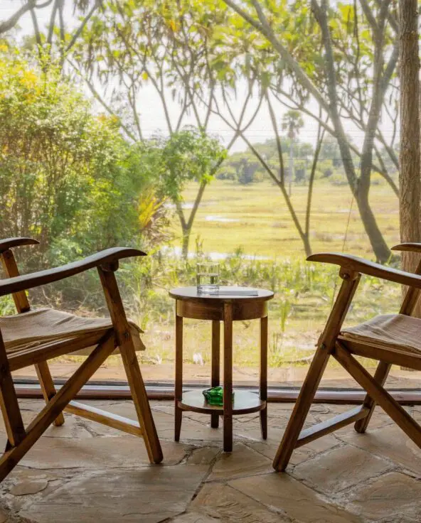 Two wooden chairs and a small table on a deck facing a green riverside view through trees at Roho ya Selous Camp