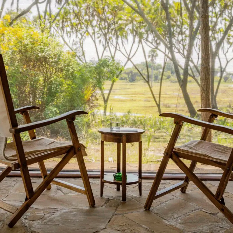 Two wooden chairs and a small table on a deck facing a green riverside view through trees at Roho ya Selous Camp