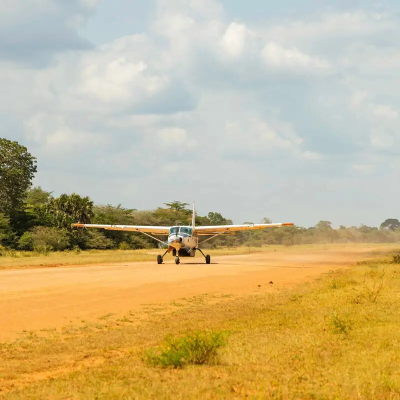 A small propeller plane taxiing on a dusty airstrip surrounded by grassy savannah