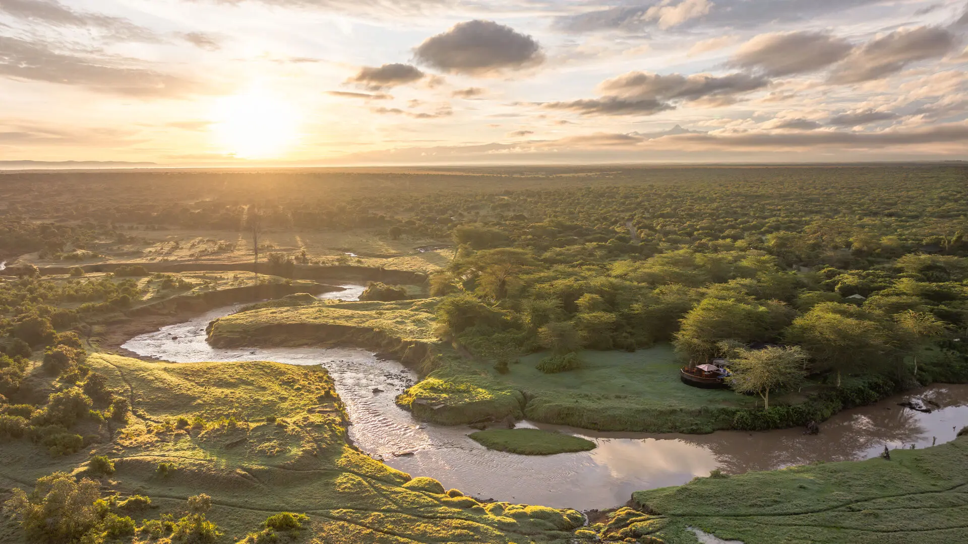 Aerial view of Ol Pejeta Bush Camp nestled in lush green acacia woodland on a bend of a winding river at sunrise, with the vast Laikipia plateau stretching to the horizon under a dramatic cloudy sky, Kenya.