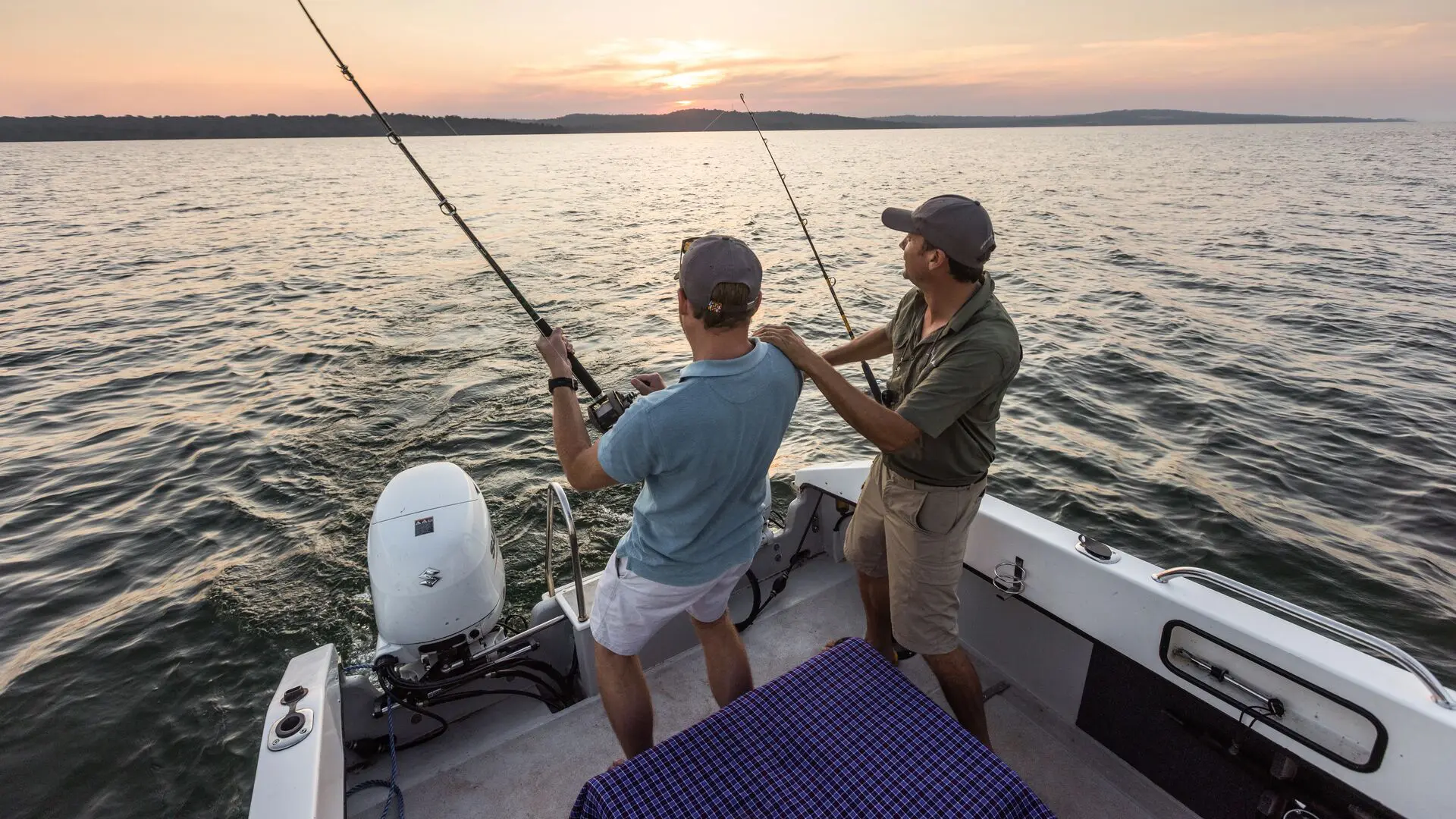 Two guests stand at the stern of a motorboat holding fishing rods over the calm waters of Lake Victoria at sunset, near Rubondo Island, Tanzania.