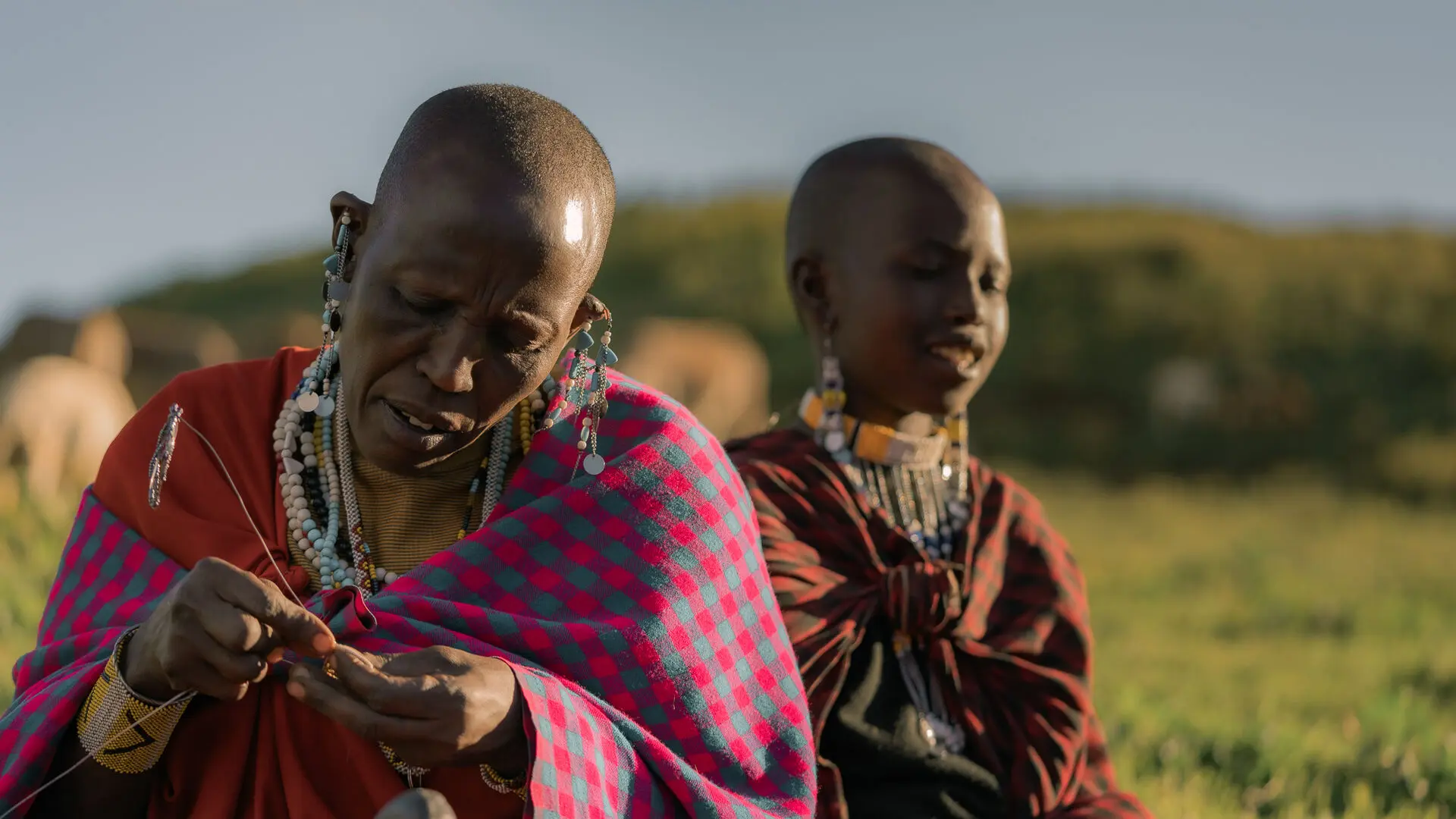 Two Maasai women wearing colourful checked shuka wraps and traditional beaded jewellery sit outdoors in soft golden light on the Ngorongoro Highlands, one carefully threading beadwork with her hands, near The Highlands by Asilia Africa.