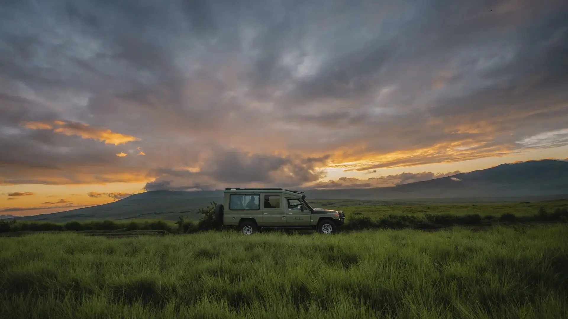 An Asilia Africa safari vehicle parked in lush green highland grass beneath a vast, dramatic sky of layered storm clouds and golden sunset light, with volcanic mountain slopes on either side, near Ngorongoro, Tanzania.