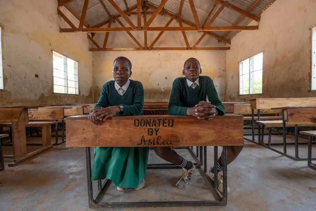 Two students in green school uniforms sit side by side at a wooden desk stamped "Donated by Asilia" in a rural classroom filled with matching desks, East Africa.