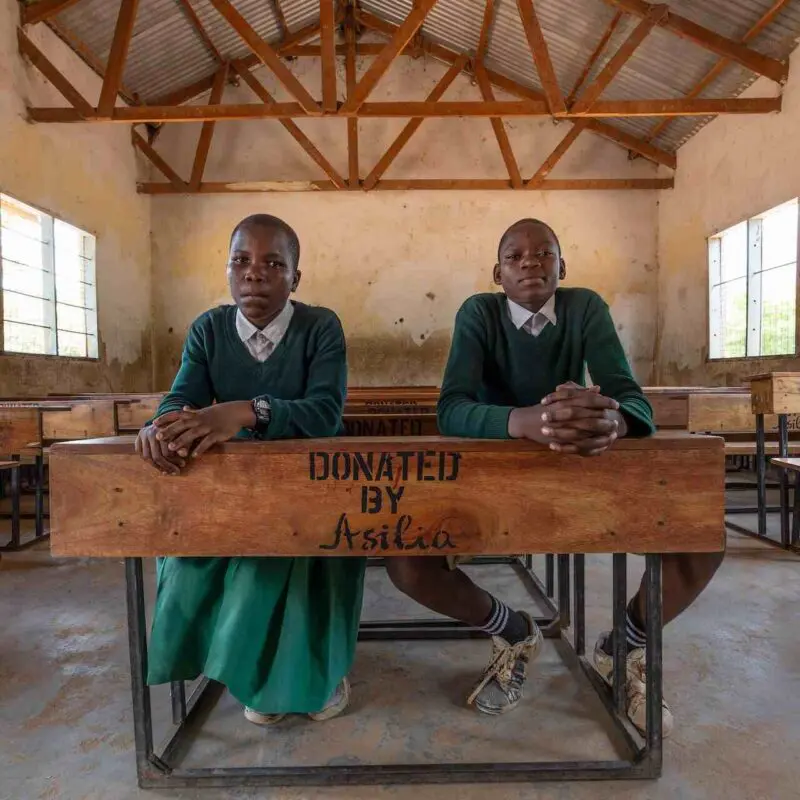 Two students in green school uniforms sit side by side at a wooden desk stamped "Donated by Asilia" in a rural classroom filled with matching desks, East Africa.