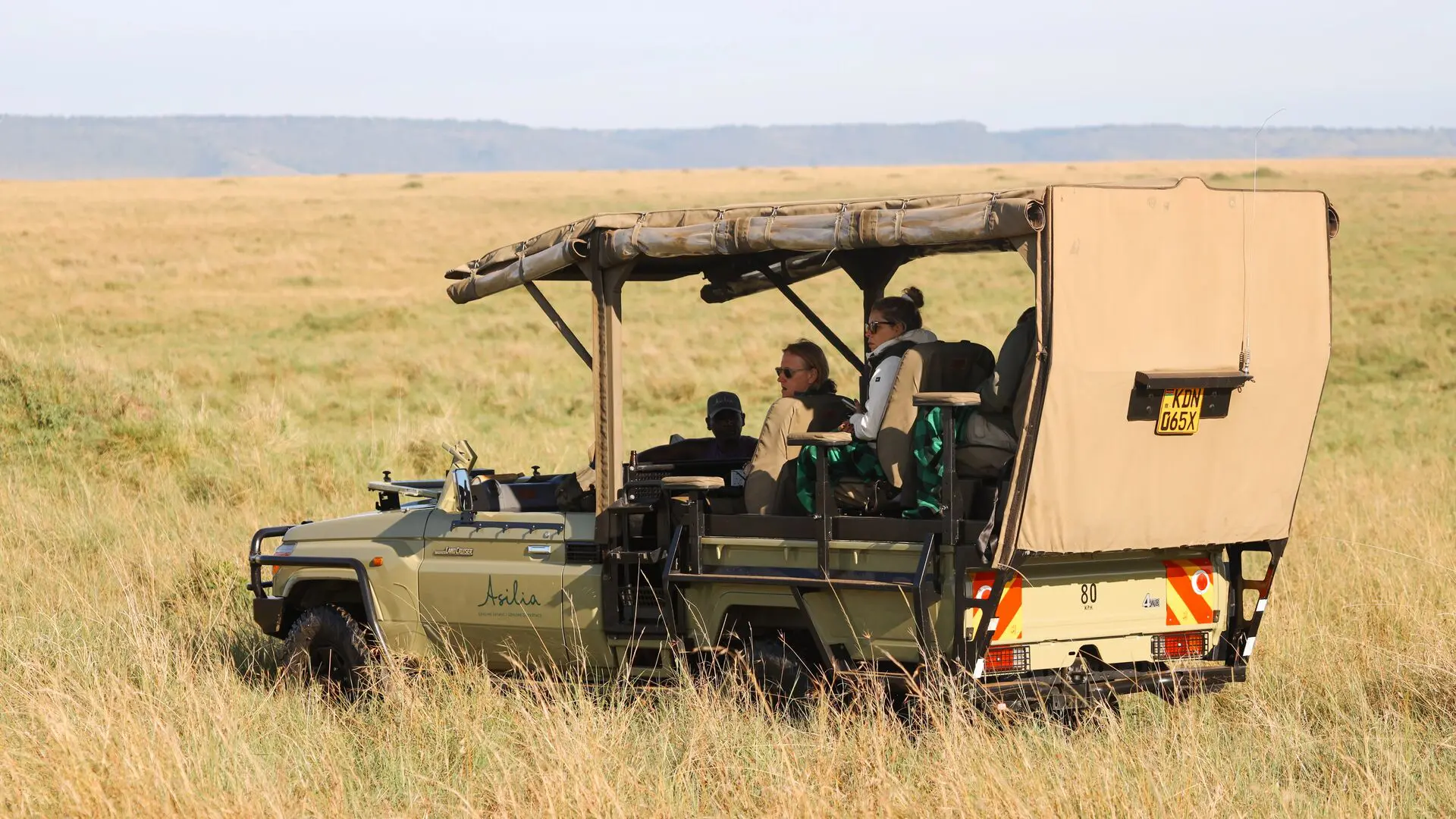 Guests seated in an open-sided Asilia Africa Land Cruiser safari vehicle scan the open Masai Mara plains, with golden grassland and the Oloololo Escarpment visible on the horizon near Rekero Camp, Kenya.