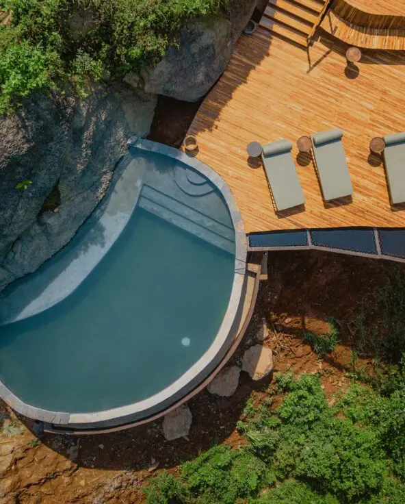 An overhead view of a curved swimming pool beside a timber deck with four loungers, surrounded by dense green vegetation and large rocks.