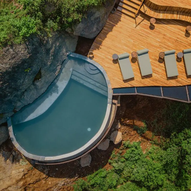 An overhead view of a curved swimming pool beside a timber deck with four loungers, surrounded by dense green vegetation and large rocks.