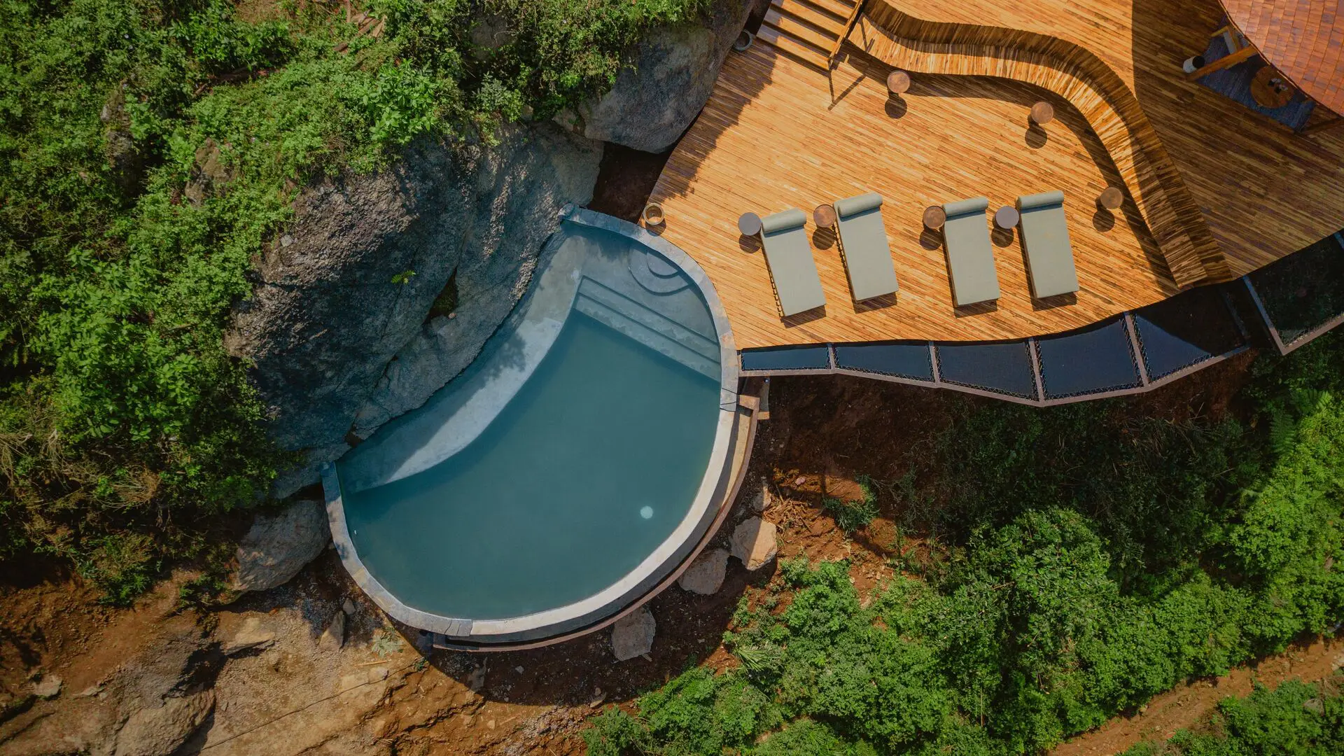 An overhead view of a curved swimming pool beside a timber deck with four loungers, surrounded by dense green vegetation and large rocks.