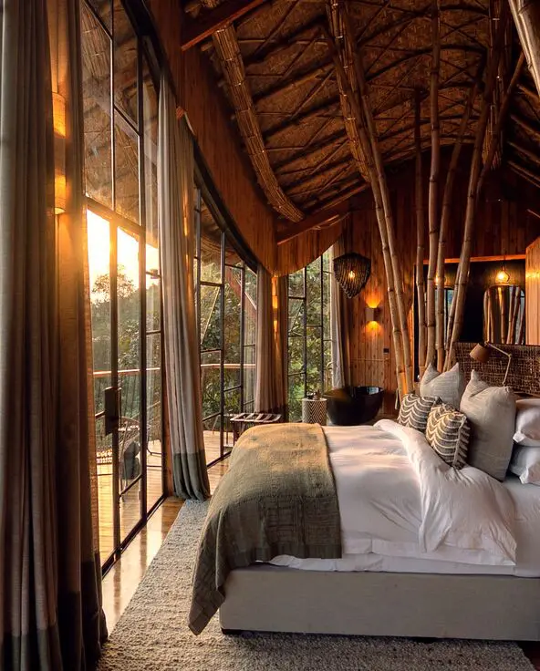 Warmly lit lodge bedroom at Erebero Hills, with a large bed, bamboo detailing and floor-to-ceiling windows opening onto a balcony and forested hills.