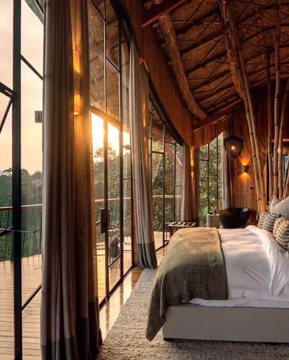 Warmly lit lodge bedroom at Erebero Hills, with a large bed, bamboo detailing and floor-to-ceiling windows opening onto a balcony and forested hills.
