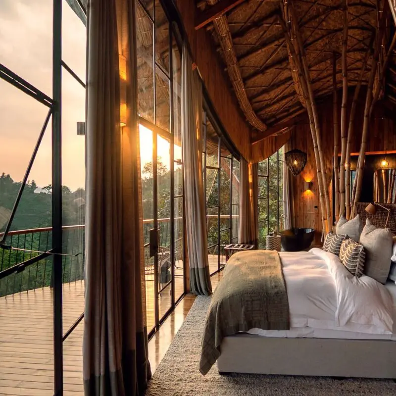 Warmly lit lodge bedroom at Erebero Hills, with a large bed, bamboo detailing and floor-to-ceiling windows opening onto a balcony and forested hills.