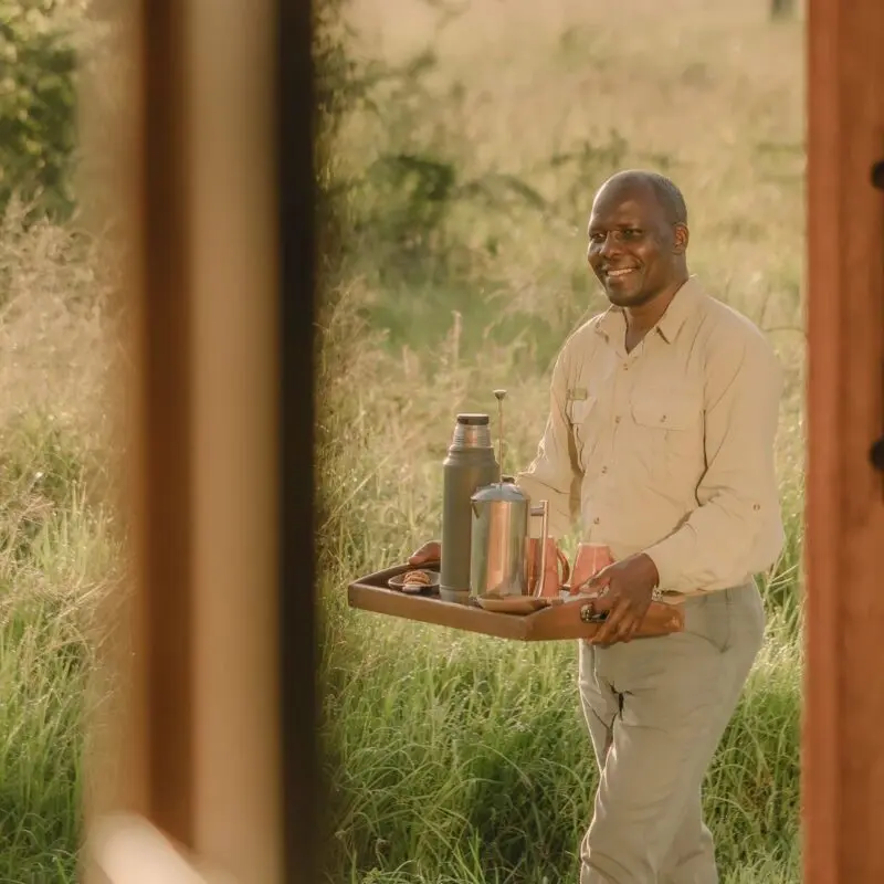 Asilia staff member carrying a tray with coffee through the grass at Naboisho Camp, Kenya.