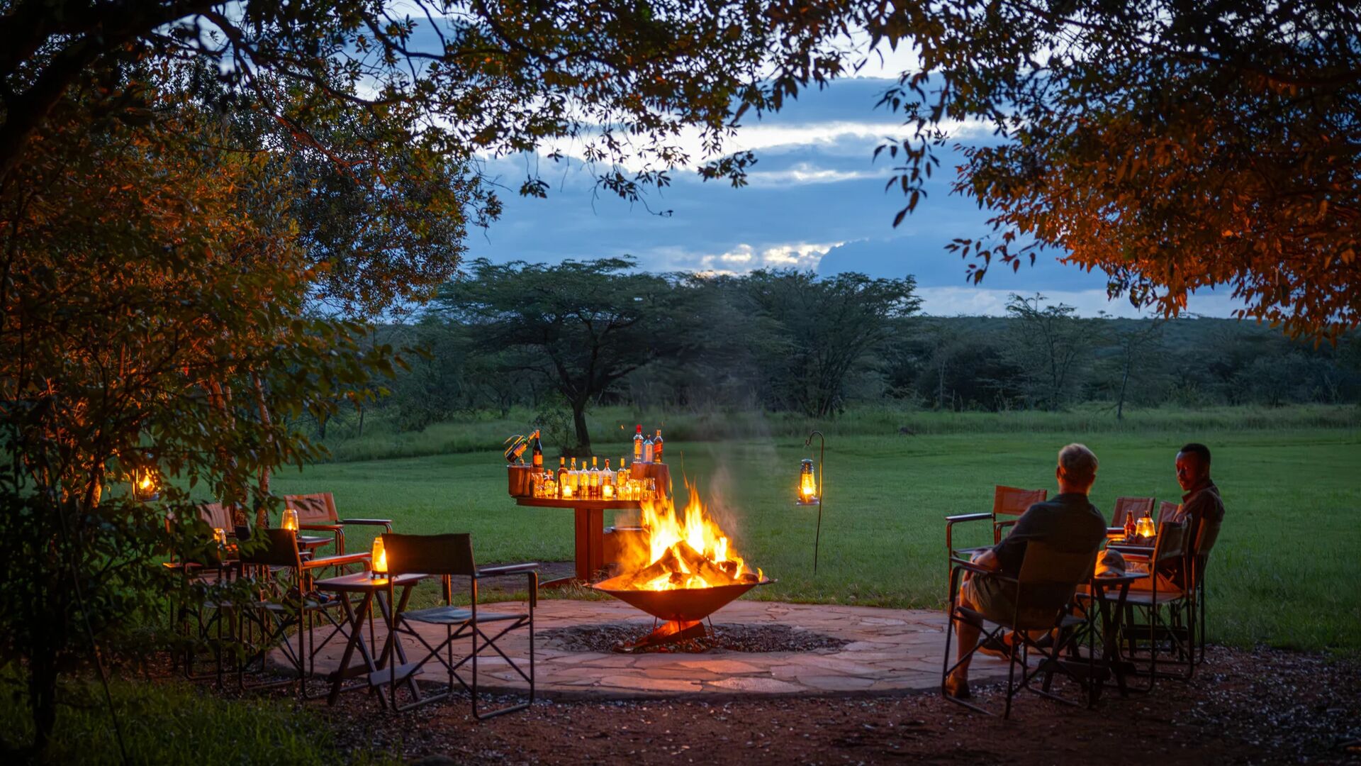 Guests of Encounter Mara Camp seated around an open campfire at dusk, enjoying drinks and lantern light overlooking open savannah and trees.