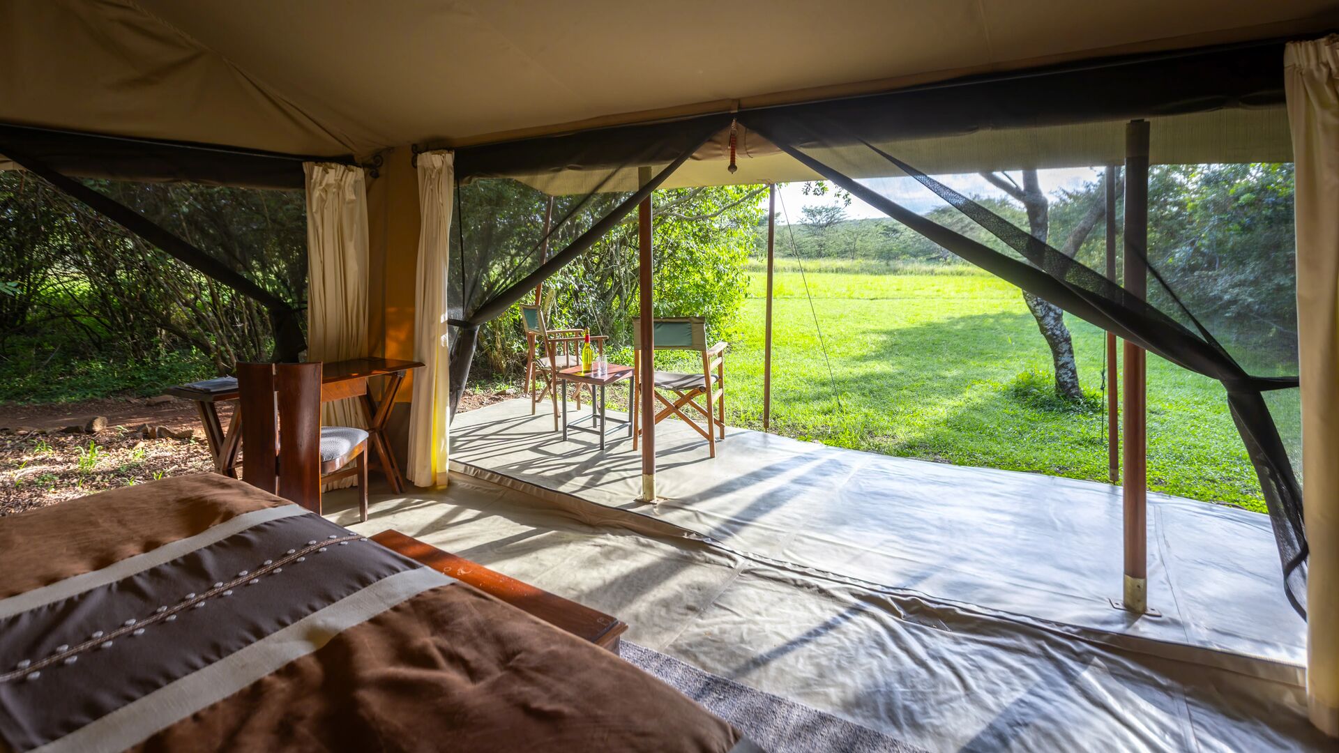 Interior of a safari tent with bed and open front leading to a grassy outdoor seating area