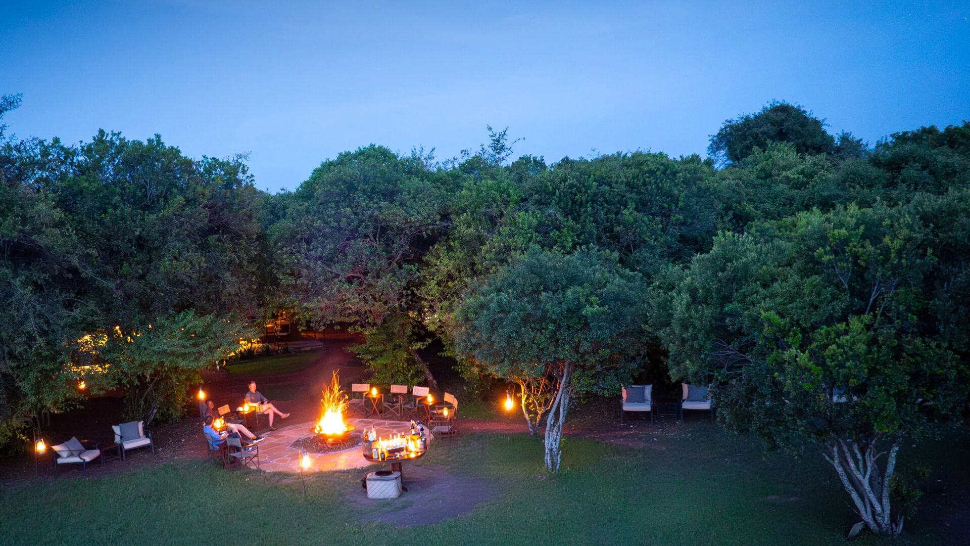 Aerial view of a safari camp at night with a central fire pit, lantern-lit seating, and surrounding trees under a deep blue sky.