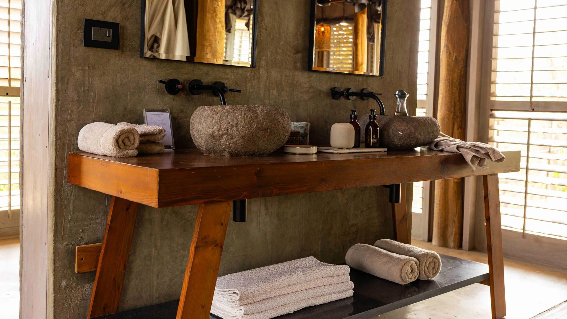 Jabali Private House bathroom with a wooden vanity, stone basin sinks, neatly folded towels, and natural light filtering through shuttered windows.