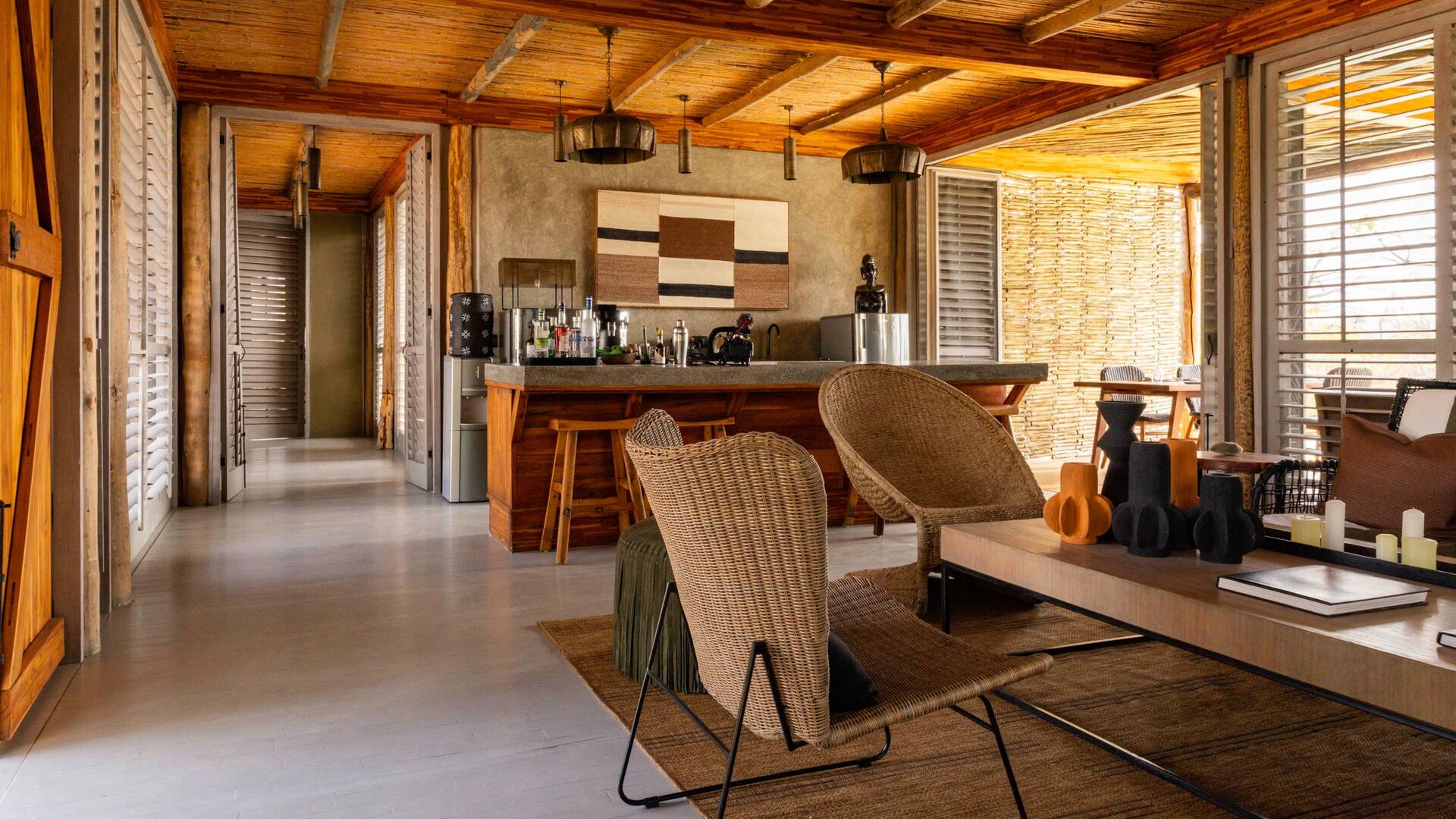 Jabali Private House lounge with woven chairs, a wooden bar, open shutters, and natural light filtering through, set within a safari lodge environment.