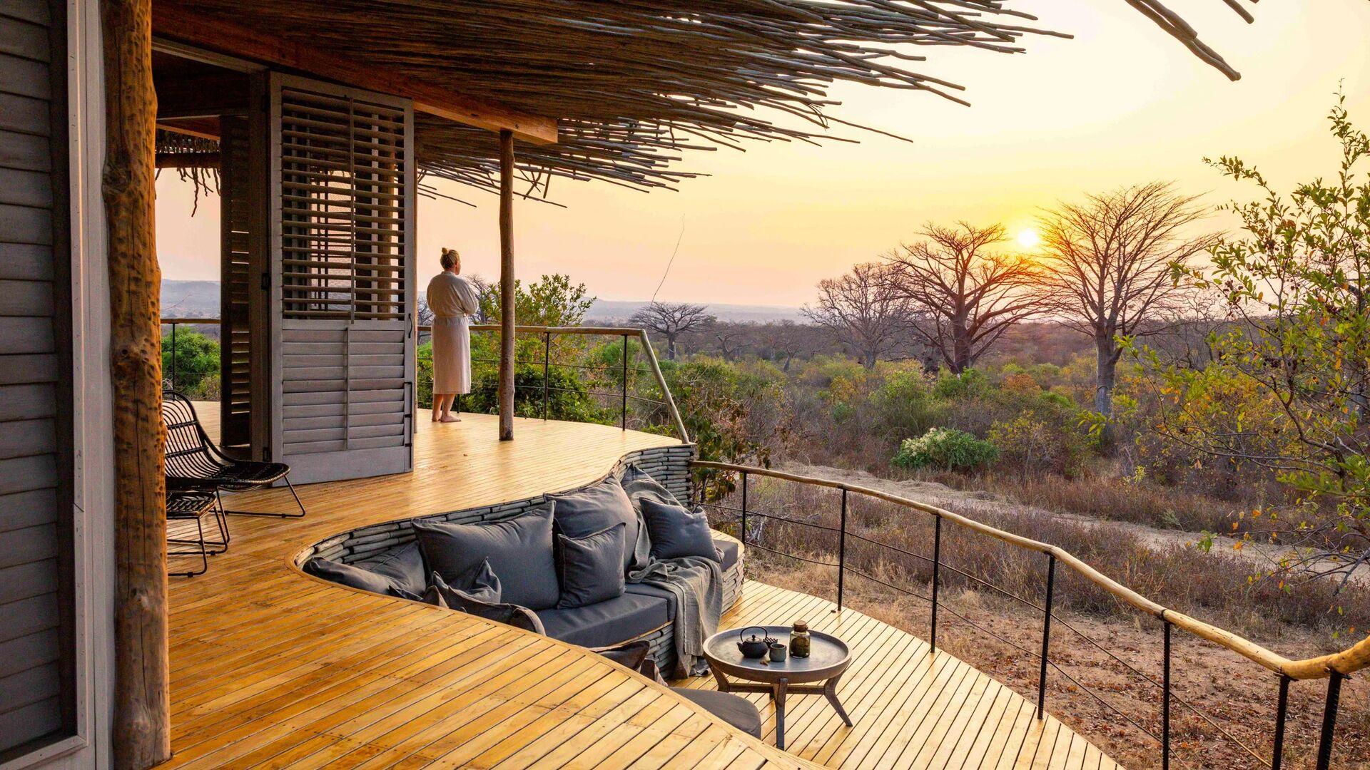 A guest stands on a private wooden deck at sunset, overlooking open woodland and baobab trees, with built-in seating and soft furnishings facing the view.
