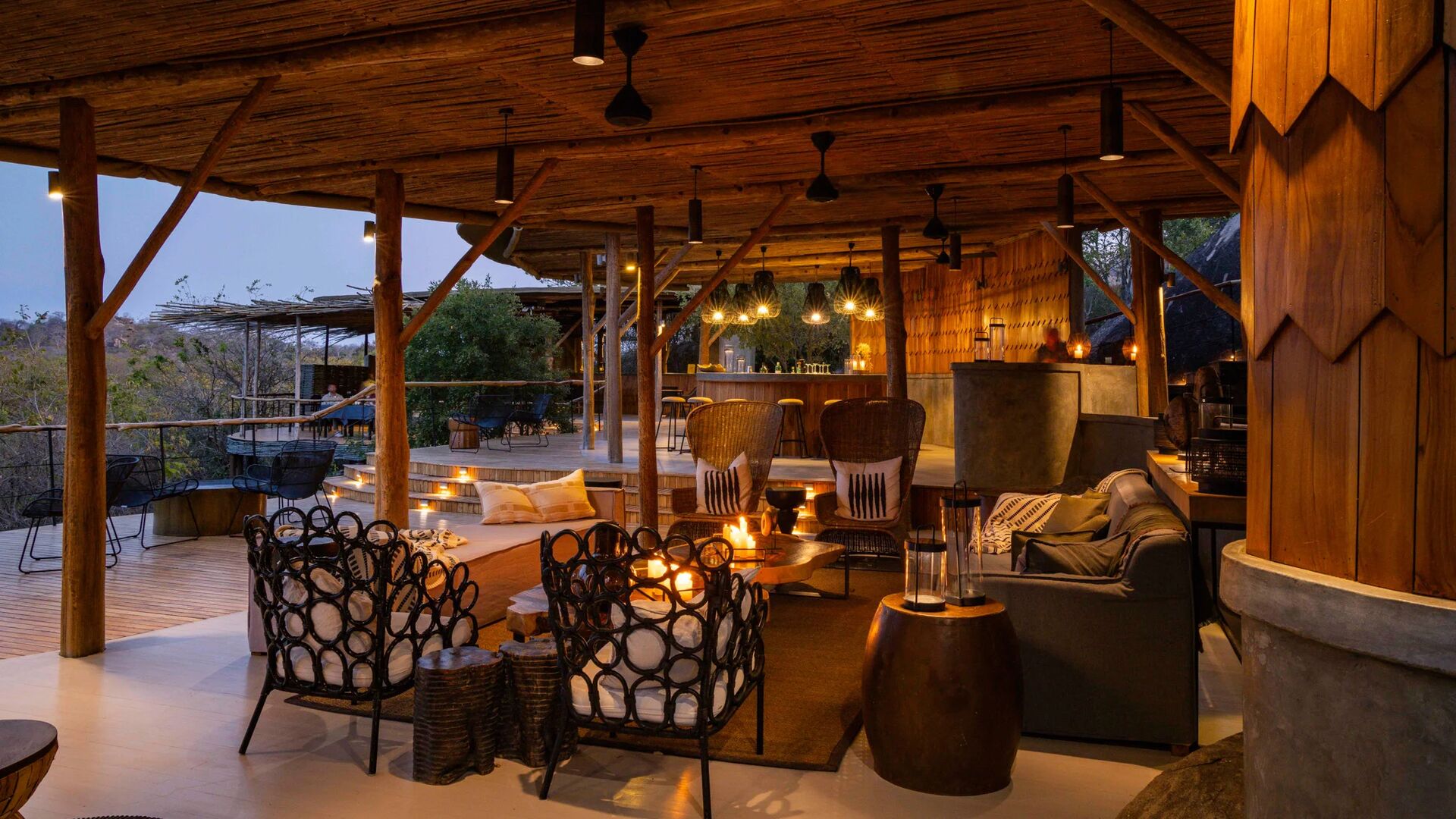 Jabali's open-air safari lodge lounge and bar at dusk, with wooden beams, warm lantern lighting, comfortable seating, and views over the surrounding bush.