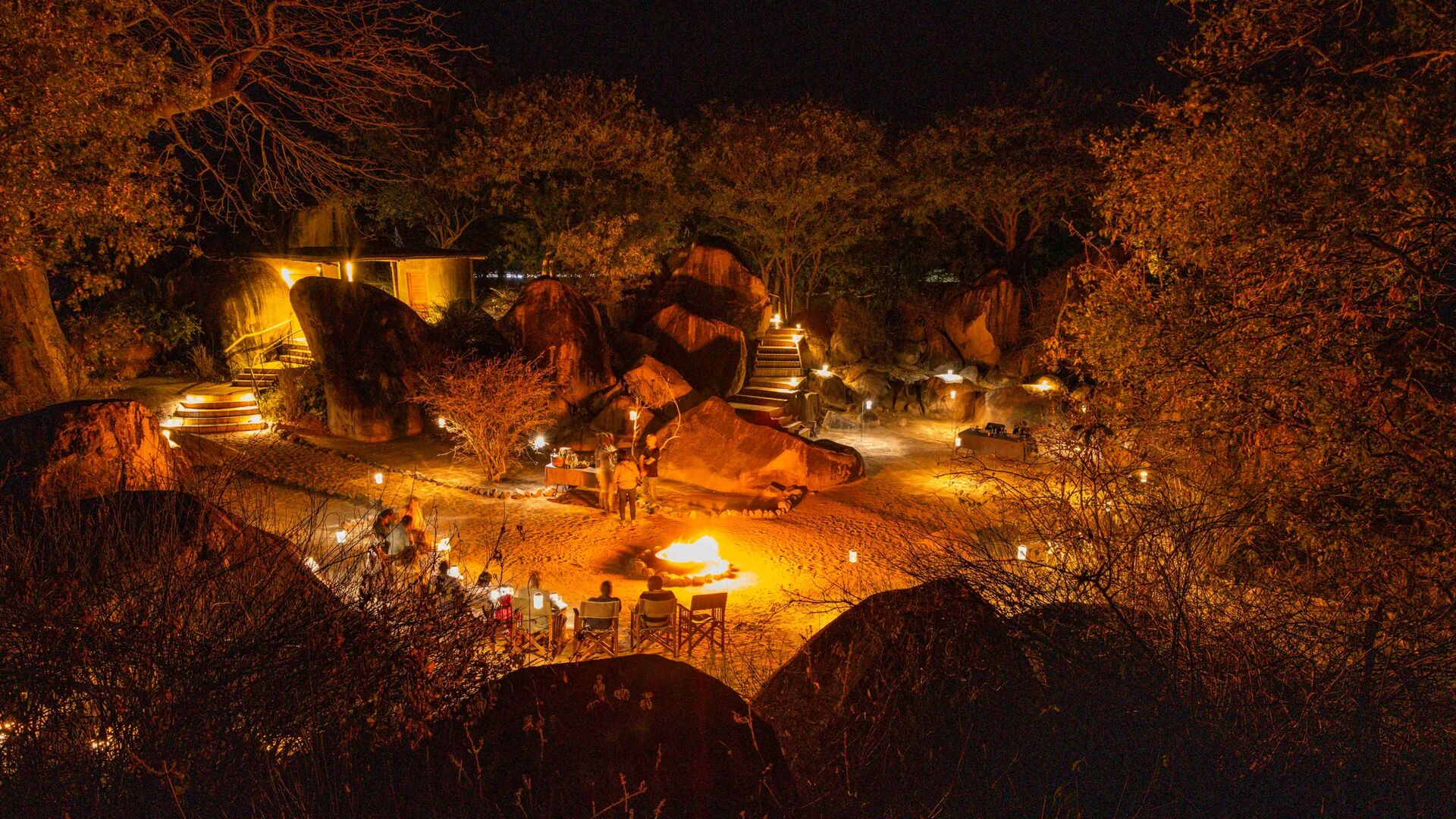Guests gather around a fire pit at night, surrounded by softly lit boulders and trees, with lanterns illuminating an open-air lodge setting beneath the stars