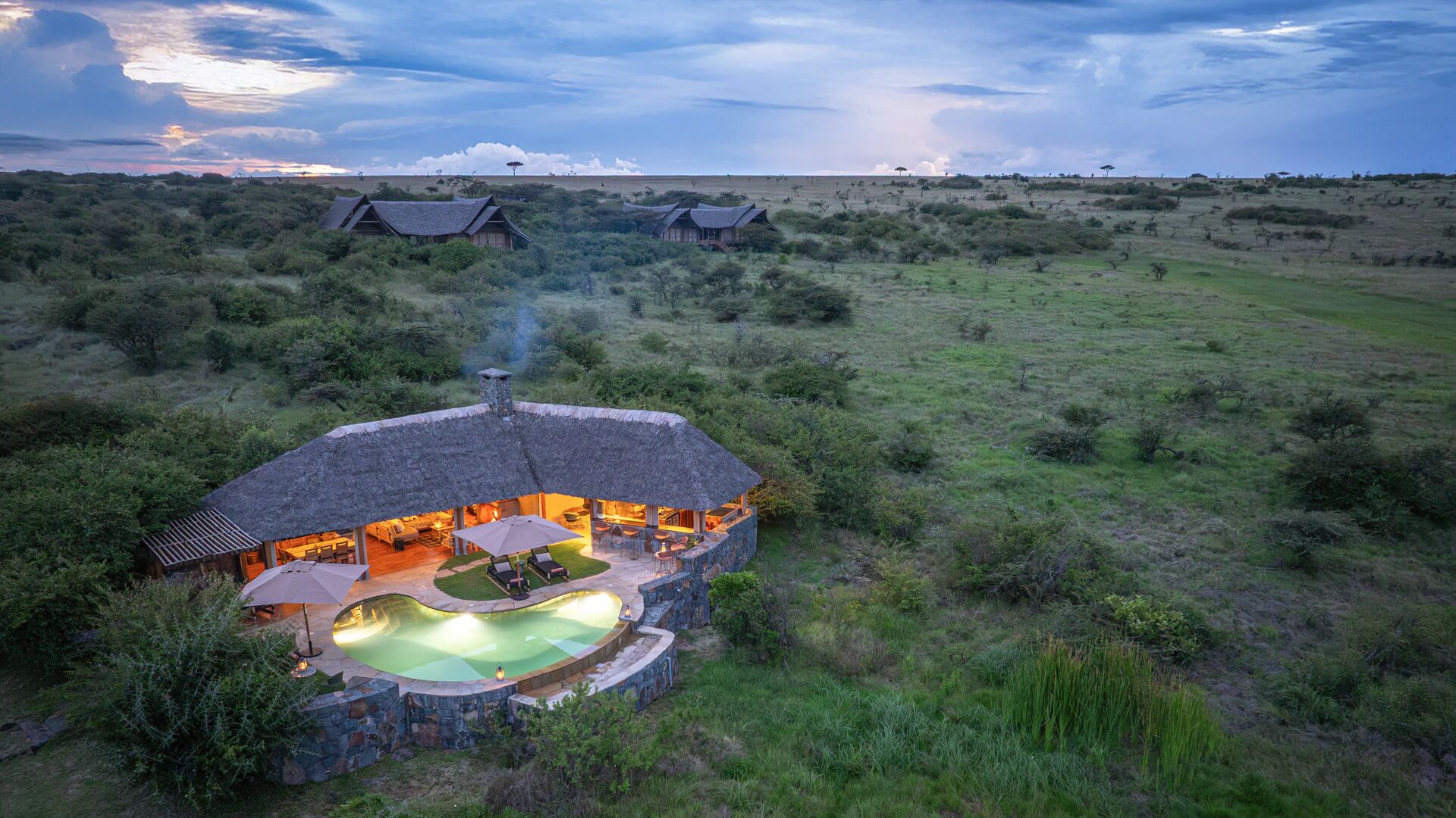 Aerial view of Naboisho Camp at dusk, featuring a lit main lodge with swimming pool, set within expansive green savannah under a dramatic evening sky.