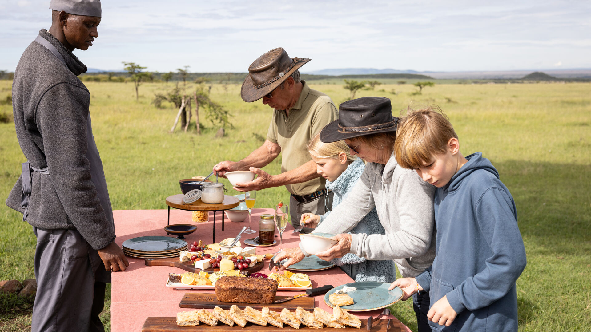 Guests and children serving themselves food at a bush breakfast table set in open grassland
