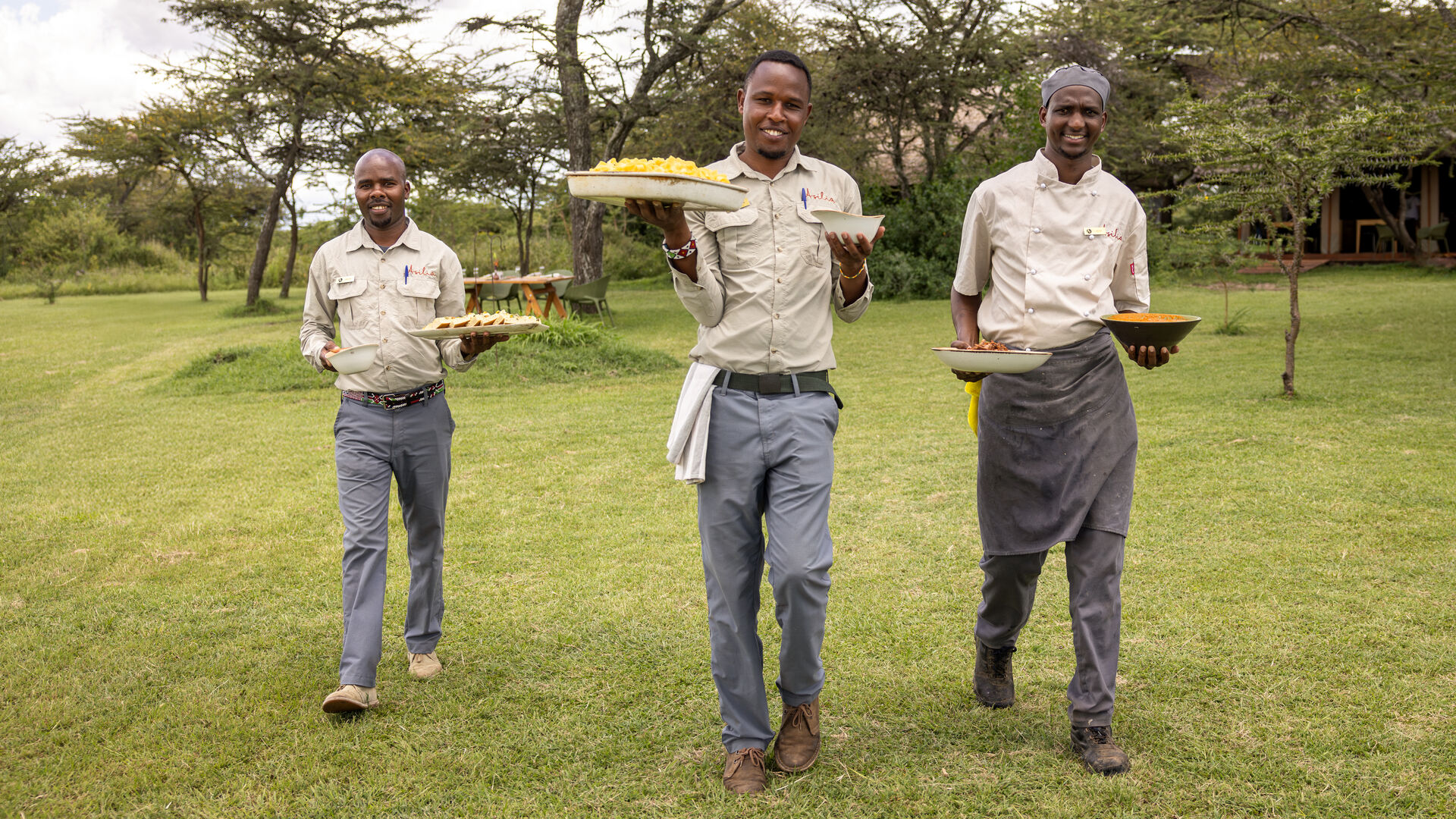 Three safari staff members walking across grass carrying plates of food toward guests