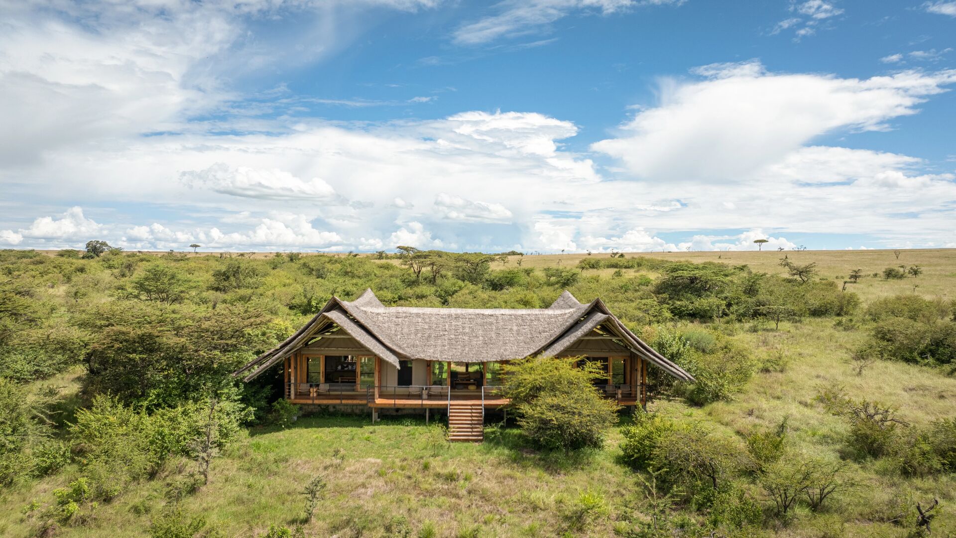 A standalone luxury family suite at Naboisho Camp with a thatched roof, raised deck, and steps, surrounded by green savannah and scattered acacia trees under a bright sky.