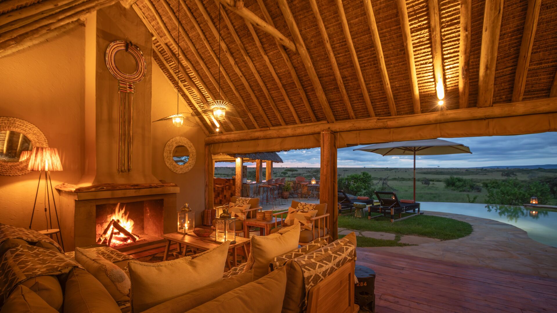 Naboisho Camp's lounge featuring a roaring fireplace, comfortable seating, wooden beams, and a deck overlooking the savannah and pool at dusk.