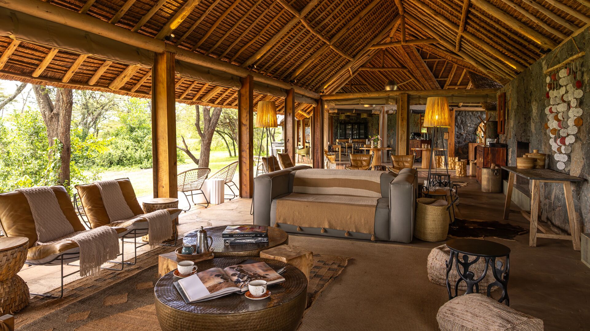 Naboisho Camp's open-sided lounge with comfortable seating, wooden beams, woven décor, and a bar area, opening onto greenery and shaded outdoor spaces.