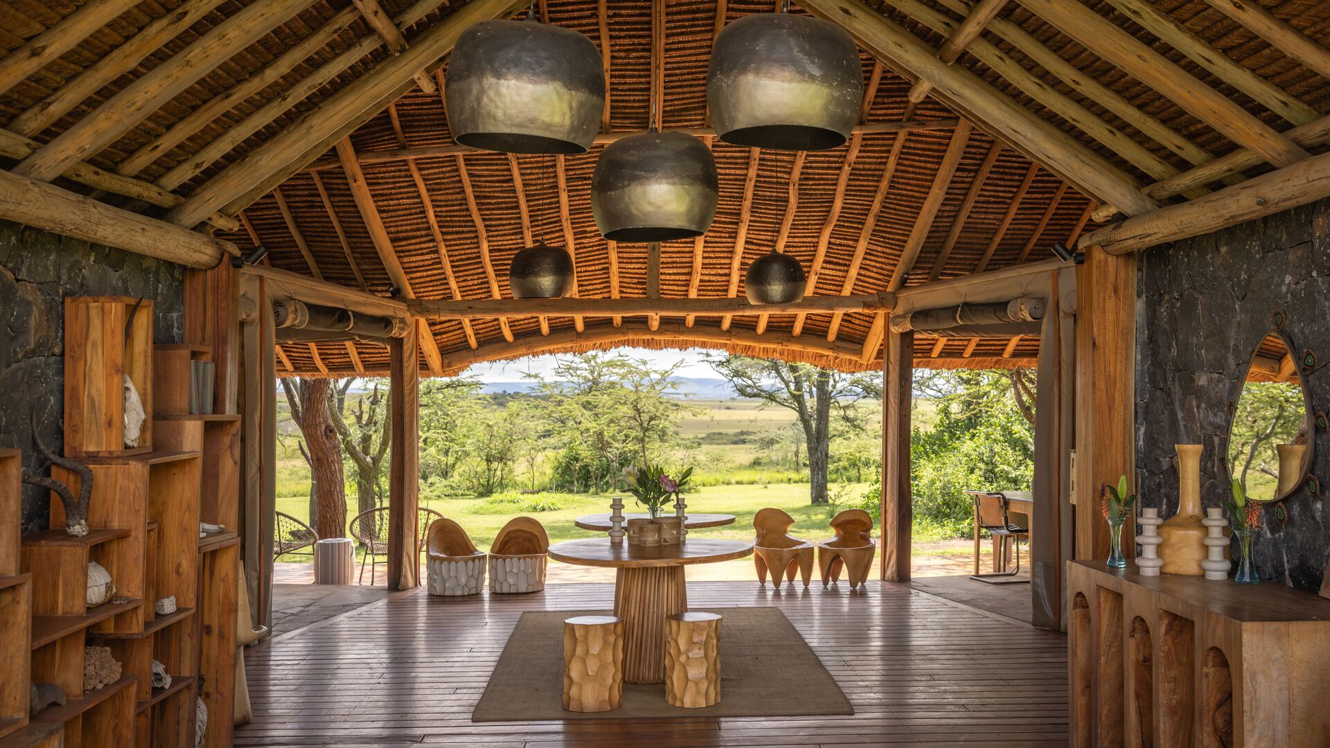 Naboisho Camp's main entrance with high thatched ceiling, wooden furnishings, sculptural seating, and open views across green savannah framed by acacia trees.