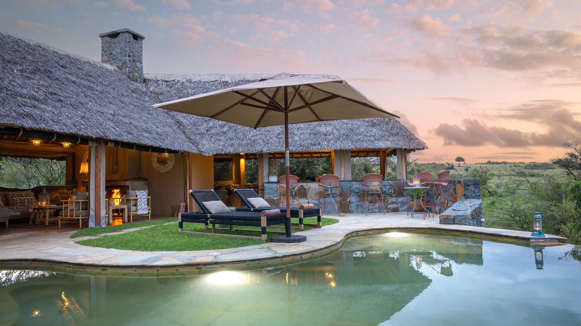Naboisho Camp's stone-edged swimming pool, shaded loungers, and open lounge areas, set against expansive savannah views at sunset.
