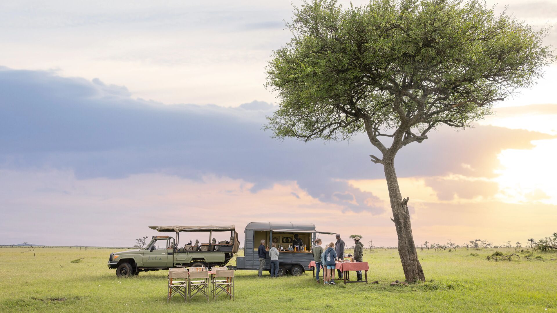 Guests gather beside a safari vehicle and mobile bar under a lone acacia tree, enjoying sundowner drinks on open grassland at sunset.