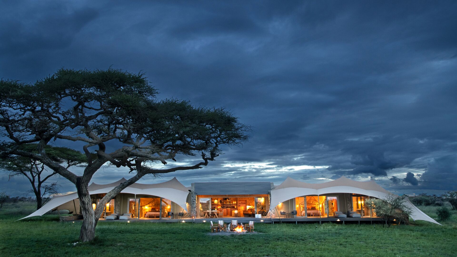 Namiri Retreat illuminated at night, with canvas tents and a central lounge glowing warmly beneath dark, dramatic clouds, set on open grassland beside an acacia tree.