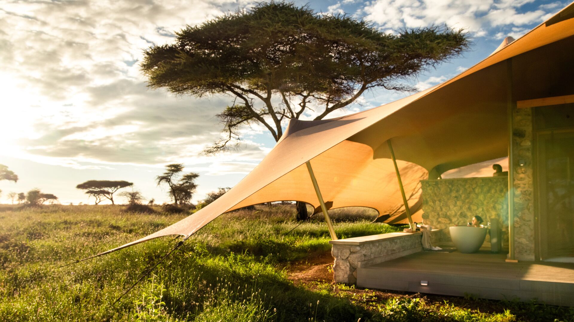 The Namiri Retreats feature an outdoor bathtub beneath a canvas canopy, set on a private deck overlooking green savannah and acacia trees in warm evening light.