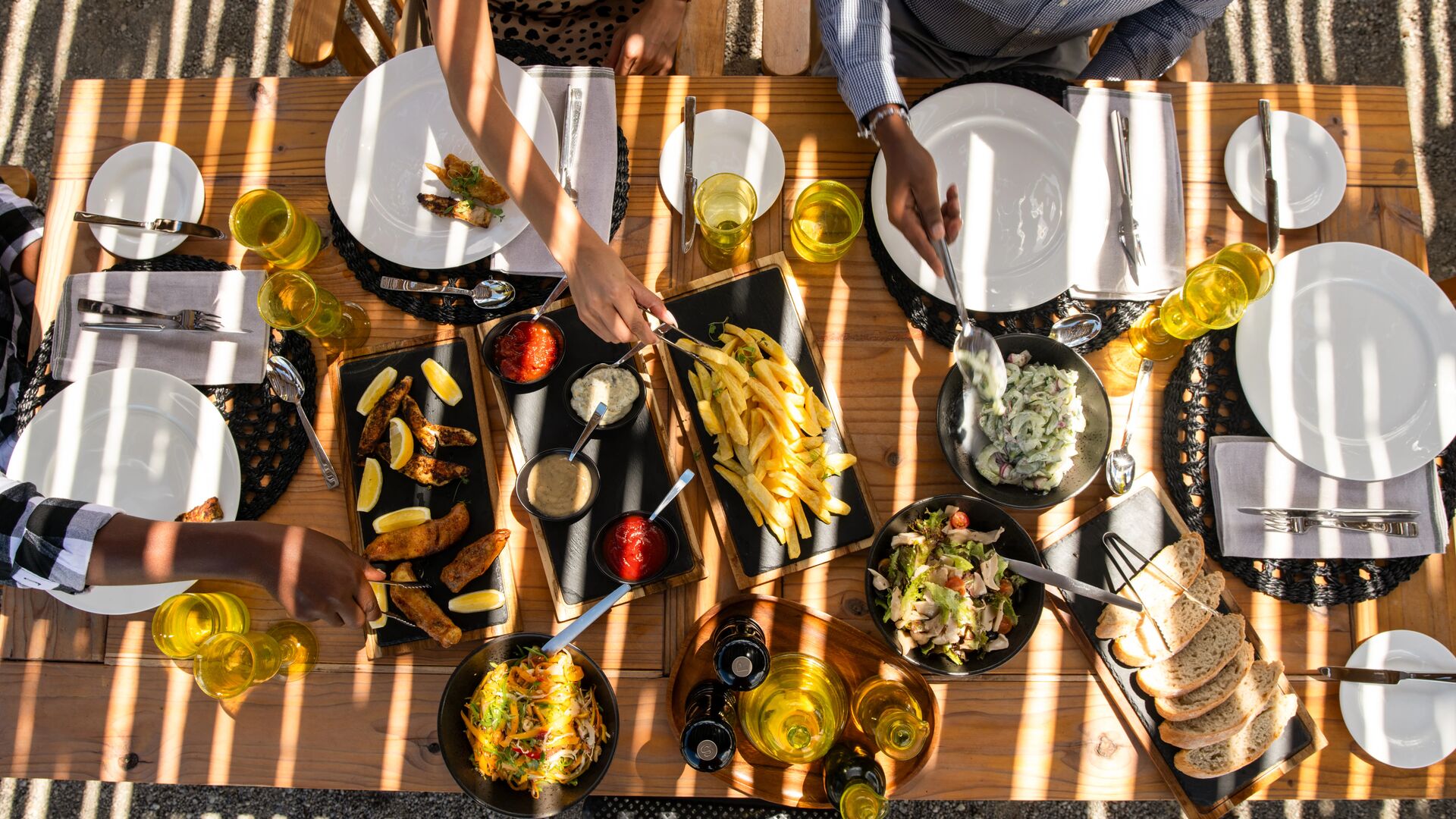 Overhead view of guests sharing a relaxed outdoor meal, with plated dishes, salads, bread, and drinks arranged on a wooden table in dappled sunlight.