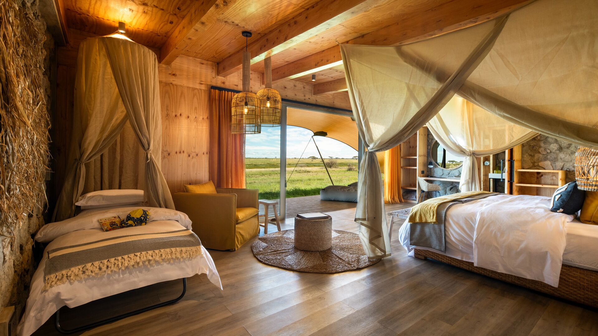 Interior of a double room with a main bed and child’s bed, soft draped mosquito nets, warm wooden finishes, and open doors leading to a deck with sweeping savannah views.