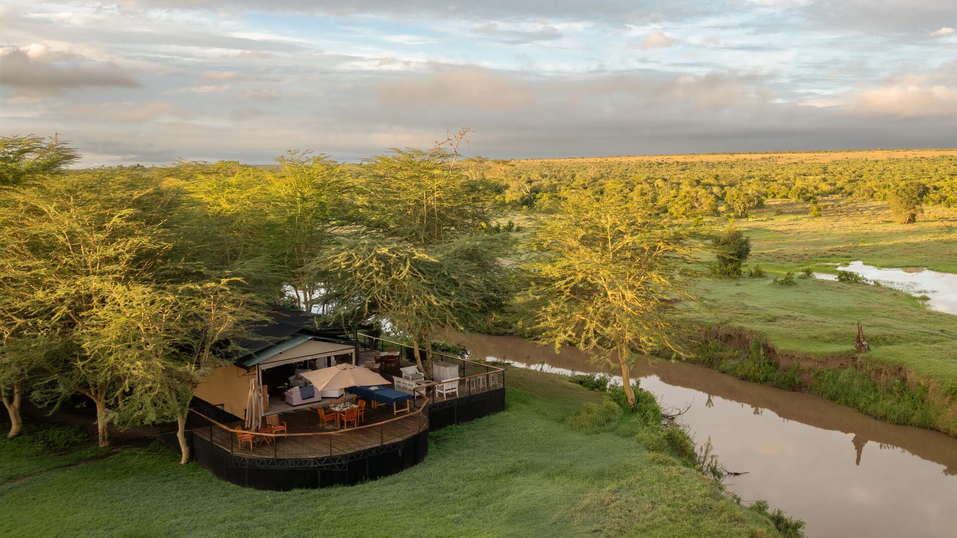 An aerial view of Ol Pejeta Safari camp set beside a winding river, with wooden decks nestled among trees and expansive green savannah stretching to the horizon under a soft, clouded sky.